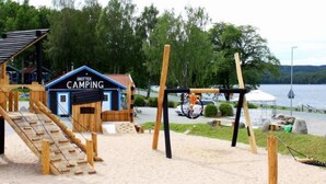 Uma estrutura de madeira para brincar com escorrega e balanços, um pequeno prédio azul com a inscrição "SKOTTEK CAMPING" e uma área arenosa perto de um lago.