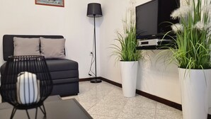 Una sala de estar moderna con un sofá, un televisor, una lámpara de pie y dos plantas grandes en macetas.