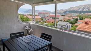 Un balcón con mesa y sillas, con vista a un pueblo costero con edificios de techo rojo y montañas al fondo.