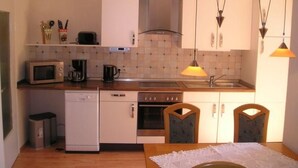 A compact kitchen with white cabinets, a microwave, a coffee maker, and a dishwasher. There are two chairs and a table with a lace tablecloth. - Haus am Reithmoor by Interhome (Großefehn)