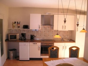 A compact kitchen with white cabinets, a microwave, a coffee maker, and a dishwasher. There are two chairs and a table with a lace tablecloth. - Haus am Reithmoor by Interhome (Großefehn)