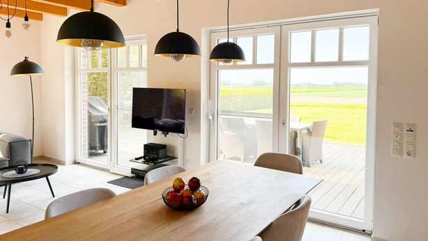 Un'area pranzo moderna con tavolo in legno, sedie e una ciotola di frutta. Si vede una televisione e un barbecue fuori dalla finestra.