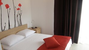 Hotellihuone, jossa on suuri sänky, valkoiset vuodevaatteet, punainen heittopeitto, yöpöytä lampun kanssa ja seinällä taideteos.
