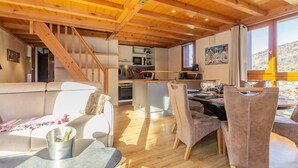 Un interior acogedor con techo de madera, comedor con sillas y mesa, cocina con electrodomésticos, y sofá con una botella de vino.