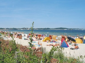 Uma praia com várias tendas e guarda-sóis coloridos, uma faixa de areia e uma vista clara do mar e colinas ao longe.