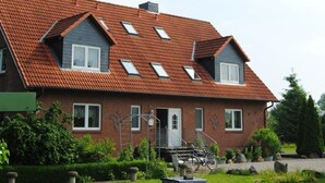 Ein zweigeschossiges Haus mit rotem Ziegeldach, zahlreichen Fenstern und einem kleinen Garten mit Steintöpfen und einem Fahrrad.