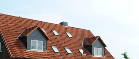 Ein zweigeschossiges Haus mit rotem Ziegeldach, zahlreichen Fenstern und einem kleinen Garten mit Steintöpfen und einem Fahrrad.