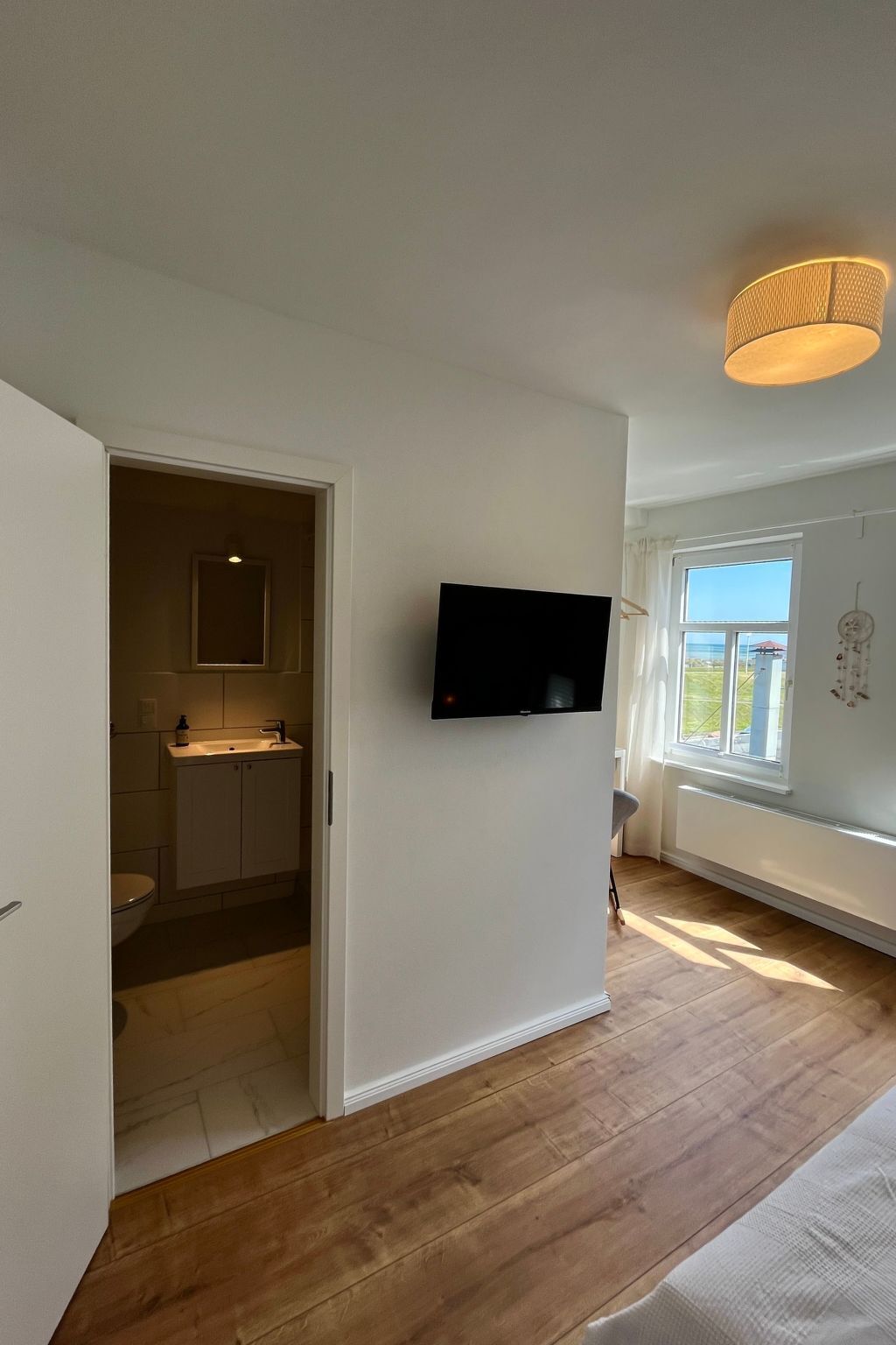 Una habitación con piso de madera, un televisor de pantalla plana montado en la pared, una ventana con cortinas y una puerta que da a un baño.