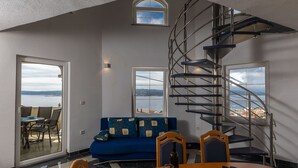 Una sala de estar moderna con un sofá azul, una mesa de comedor con sillas, una escalera de caracol y grandes ventanales con vistas al mar.