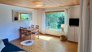 Een ruime woonkamer met houten plafond, een blauwe bank, een eettafel met stoelen, een televisie en een groot raam met gordijnen.