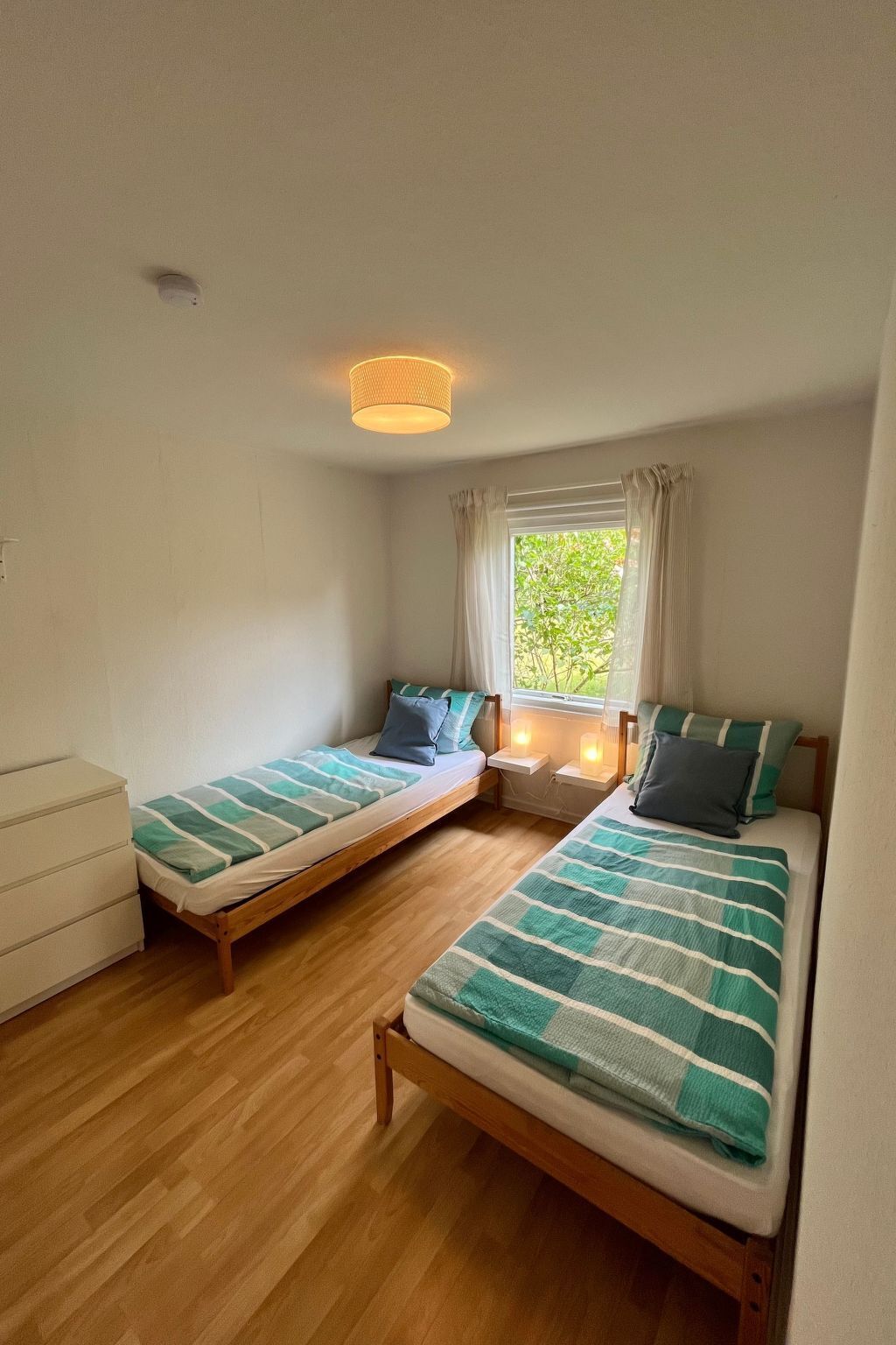 A small, well-lit room with two single beds, a wooden dresser, and a window with curtains.