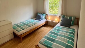 Een kleine, goed verlichte kamer met twee enkele bedden, een houten dressoir en een raam met gordijnen.