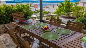 Um pátio externo com mesa de madeira posta para quatro pessoas, rodeado por cadeiras, com vista panorâmica para casas e montanhas ao fundo.
