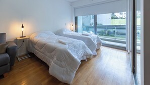 A modern bedroom with two beds, a bedside table with a lamp, and a large window with a view of the outside. - Modern Studio Apartment Full Amenities in Palermo (Buenos Aires)