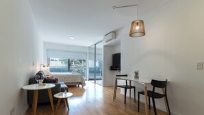 A modern, well-lit room with a bed, a dining table with chairs, a TV mounted on the wall, and a large window providing a view of the outdoors. - Modern Studio Apartment Full Amenities in Palermo (Buenos Aires)