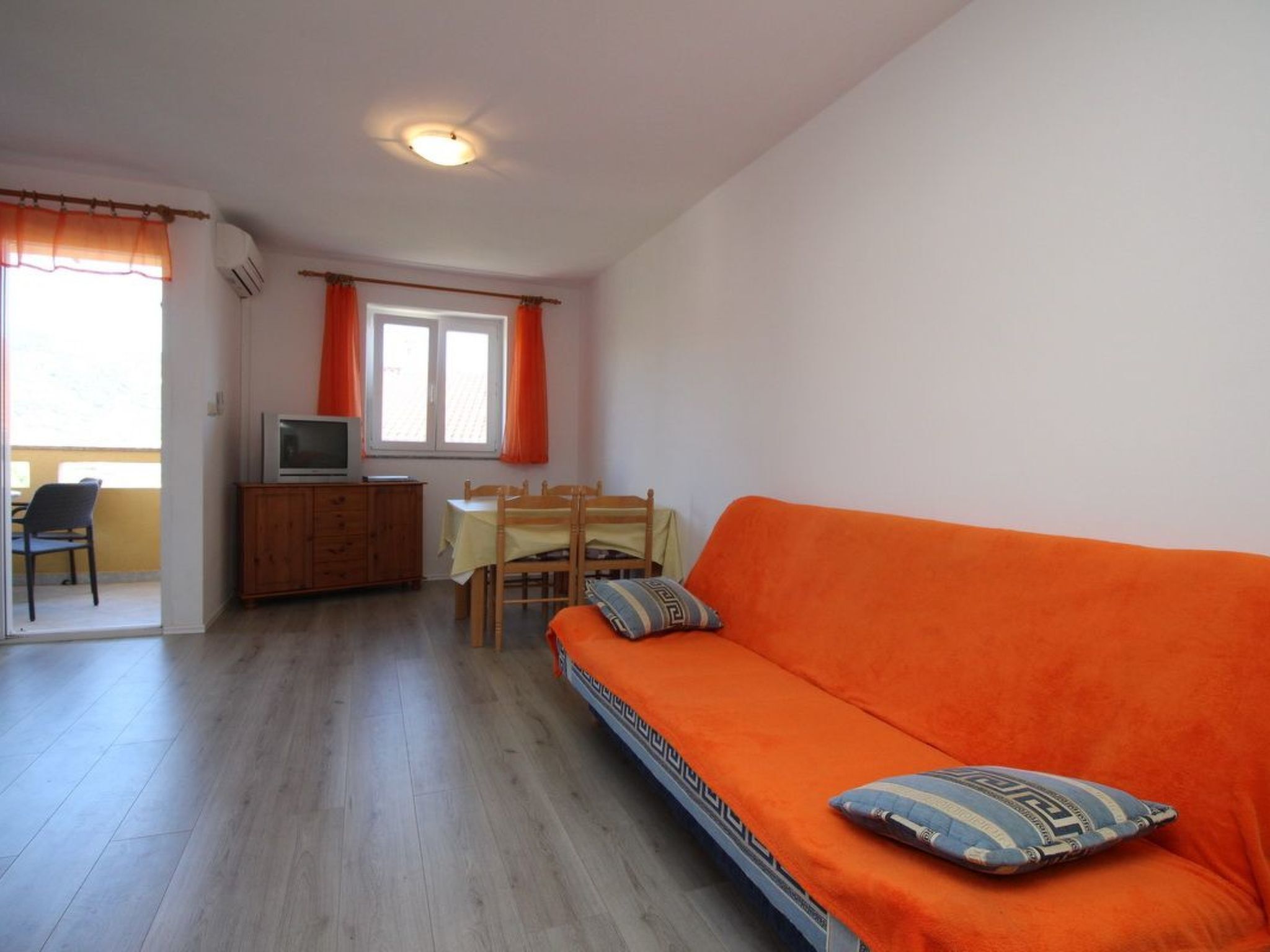 A room with a wooden floor, an orange sofa, a television on a wooden cabinet, a dining table with chairs, and a window with orange curtains.