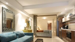Una sala de estar moderna con sofá, mesa de centro y un espejo grande. Se distingue una zona de cocina con armarios y electrodomésticos a través de un vano abierto.