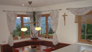 Une cuisine chaleureuse avec un banc intégré, de grandes fenêtres avec des rideaux et une croix accrochée au mur.