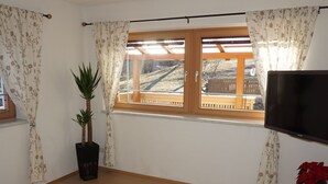 Une chambre avec un lit, une télévision, une fenêtre avec des rideaux, une plante et une vue sur une terrasse en bois à l’extérieur.