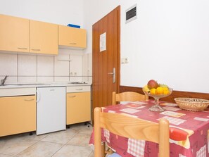 A compact kitchen with a white dishwasher, wooden cabinets, and a dining table set with a fruit bowl and a basket. - ZIC-MRAKOVCIC br.2 S2 K2 ohne Balkon by Interhome (Krk/Baška)