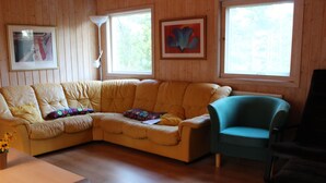 A living room with a beige sectional sofa, a teal armchair, and a black chair. There are two windows with framed artwork above them. - "Skogsmullehaus" mit Terrasse by Interhome (Loftahammar)