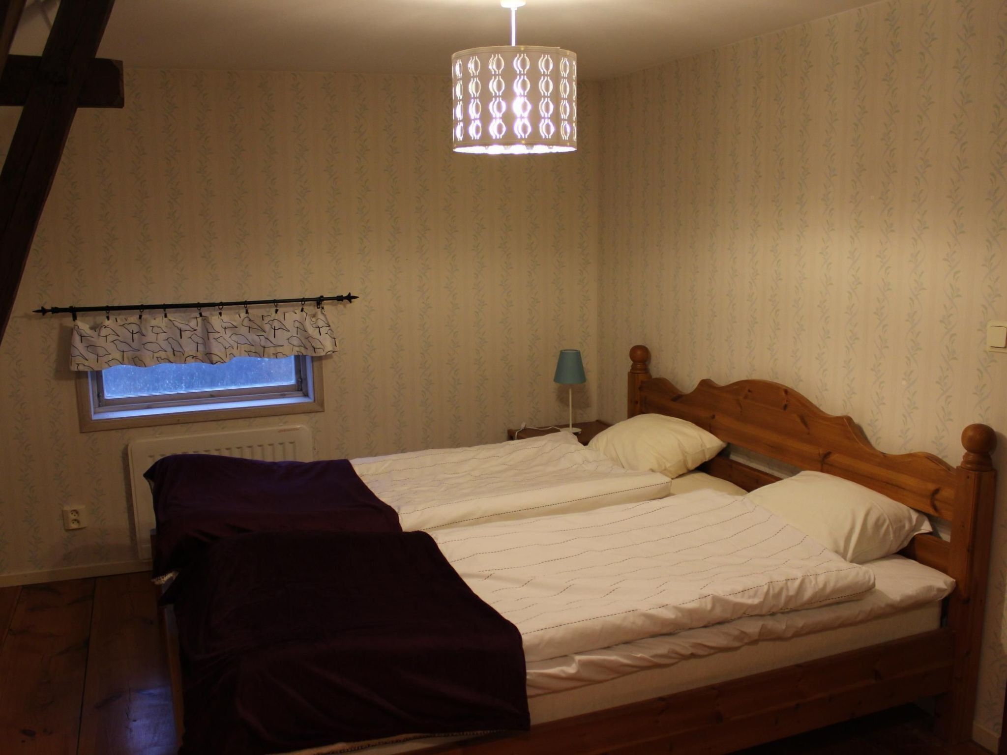 A bedroom with a wooden bed, a window with a patterned curtain, a radiator, and a hanging light fixture.