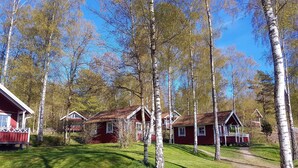 A row of red cabins with white trim, surrounded by birch trees and a clear blue sky. - Holzhaus mit überdachter Terrasse by Interhome (Bräkne-Hoby)