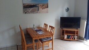 Een woonkamer met een houten eettafel, vier stoelen, een boekenkast, een flatscreen televisie en een schilderij van een strand.
