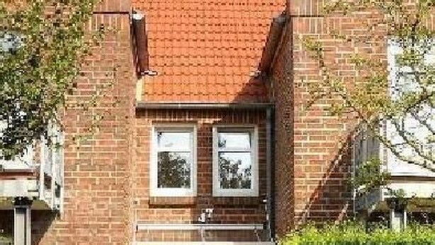 Een twee verdiepingen tellend bakstenen huis met een dak van rode tegels, een voordeur en een klein tuintje met een bankje.