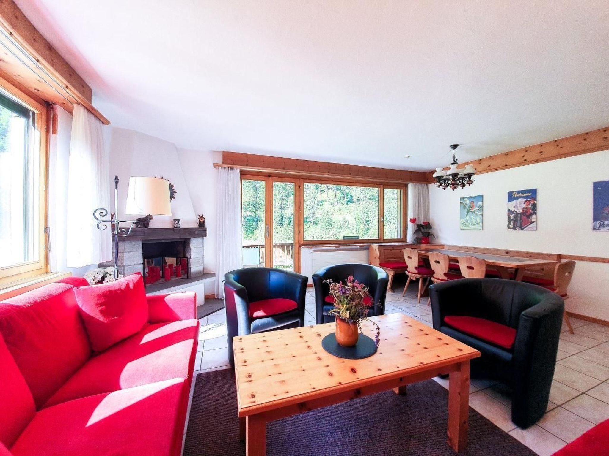 Ein gemütliches Wohnzimmer mit einer roten Couch, einem Holztisch und einem Kamin. Mehrere Stühle und ein Esstisch mit roten Kissen sind vorhanden.