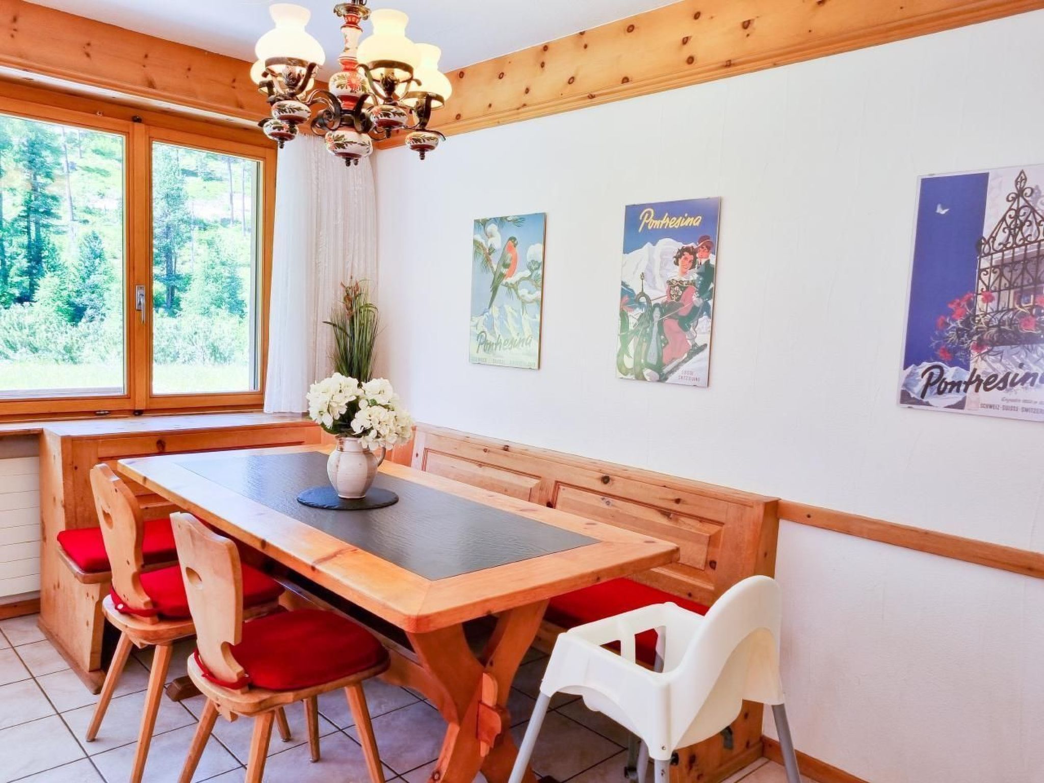 A wooden dining table with red cushioned chairs, a vase of white flowers, and a chandelier.