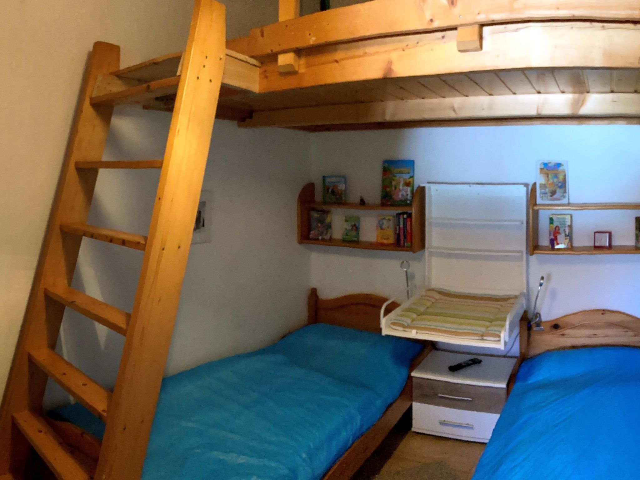 A bunk bed room with a wooden ladder, two beds with blue bedding, a small bedside table, and a shelf with books and small items.