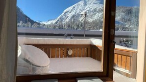 Una habitación acogedora con un banco de madera y cojines, ventanas amplias que ofrecen vistas a montañas nevadas y un lago.