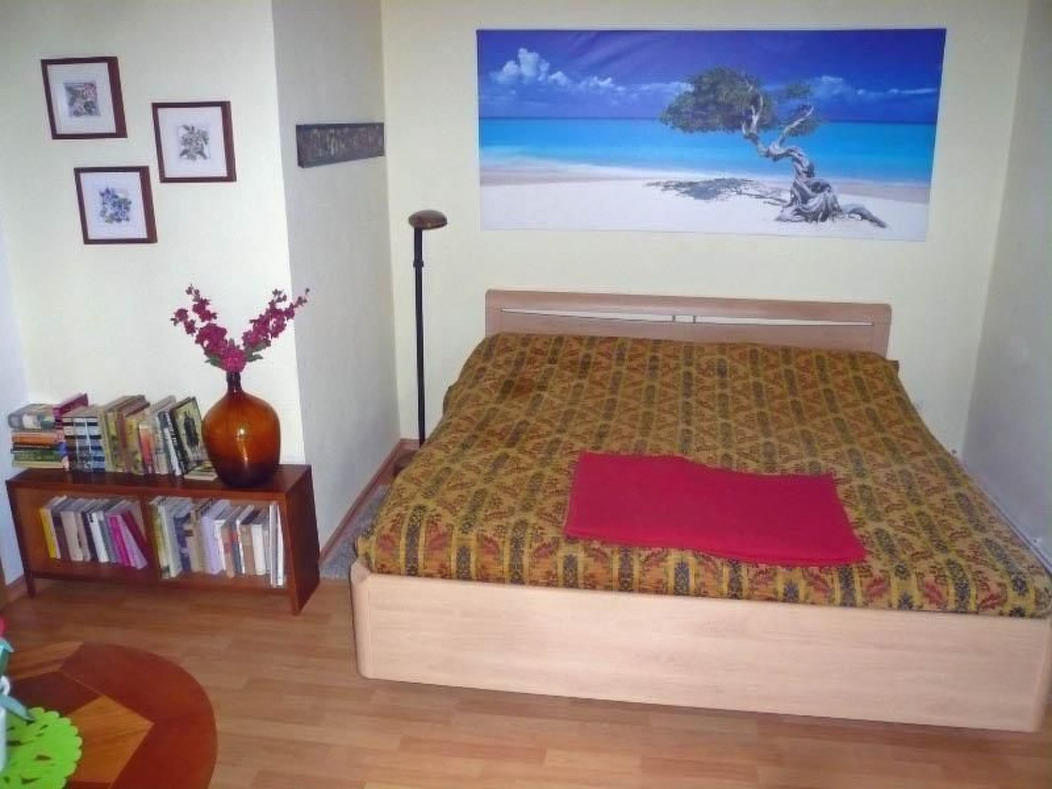 A bedroom with a bed, a bookshelf, a vase with flowers, and a painting of a beach scene.