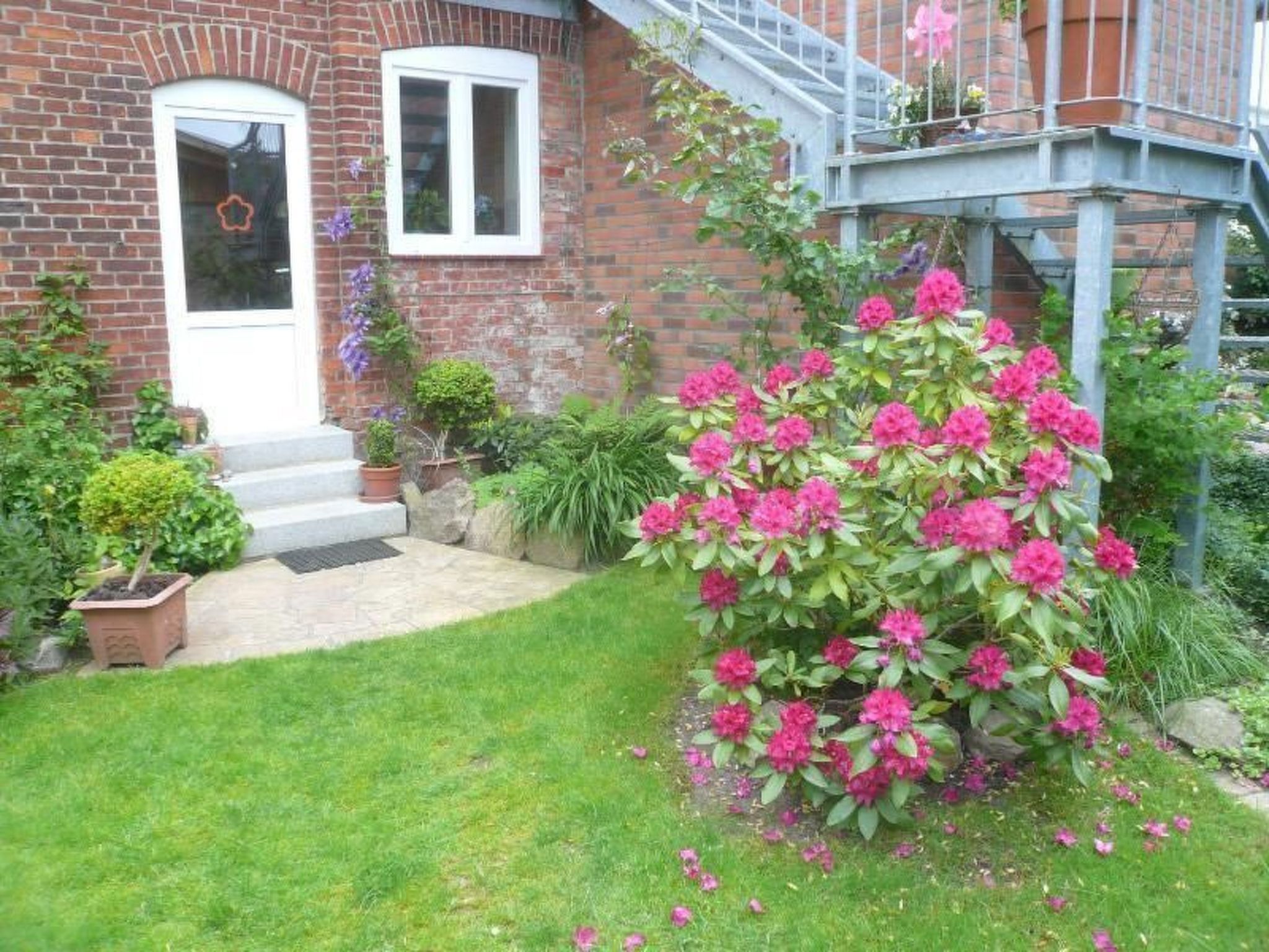 Una casa in mattoni con una porta d'ingresso, due finestre, un piccolo giardino con fiori rosa e una scalinata blu.