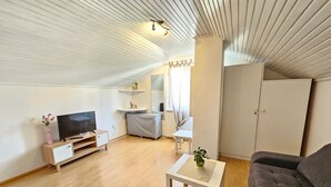 Ein geräumiges Wohnzimmer mit Flachbildfernseher, Holzboden, einem grauen Sofa, einem kleinen weißen Tisch und einem Kleiderschrank.