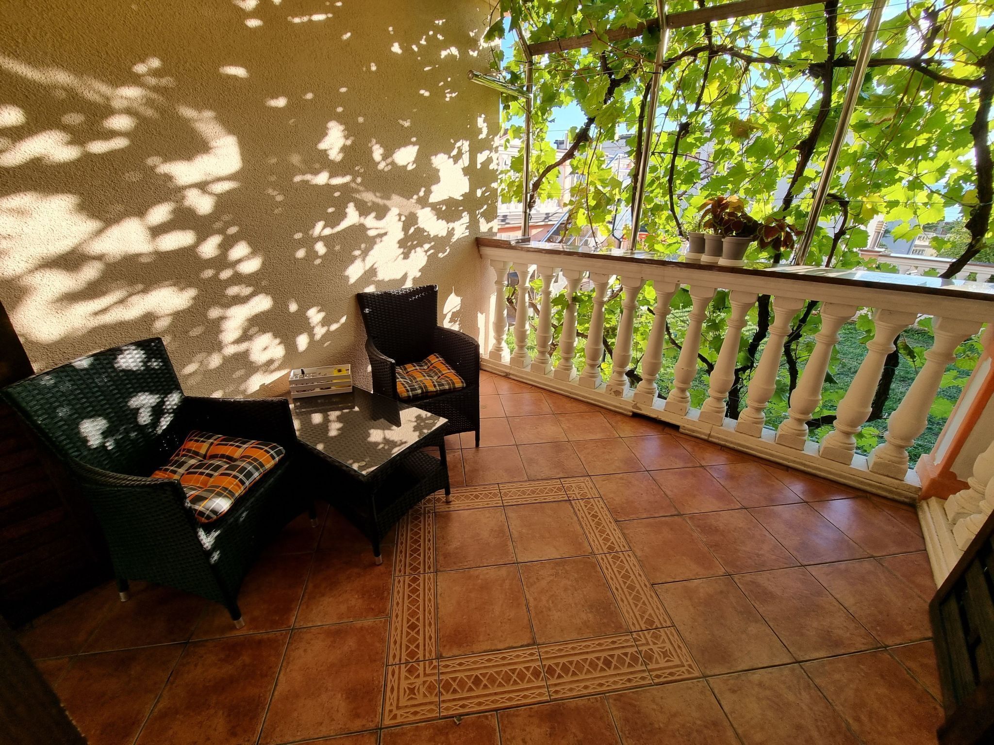 Uma área de pátio com duas cadeiras de vime, uma pequena mesa, cerca decorativa branca e folhagem verde ao fundo.
