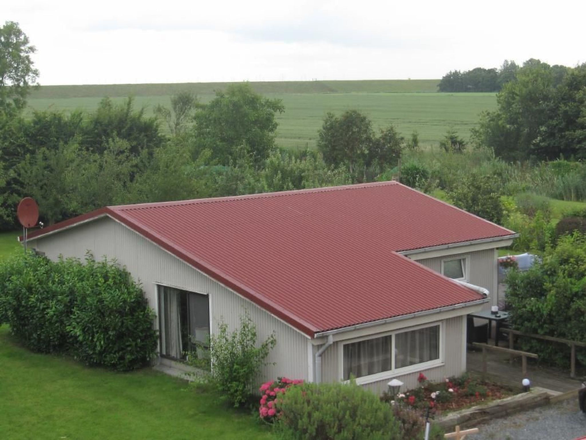 A single-story house with a red roof, surrounded by greenery and a large field in the background.