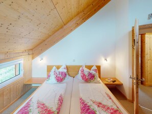 A bedroom with a wooden ceiling, a bed with floral bedding, a bedside table with a book, and a window with a view of greenery. - Klippitzhimmel in Klippitztörl by Interhome (Bad St. Leonhard im Lavanttal)