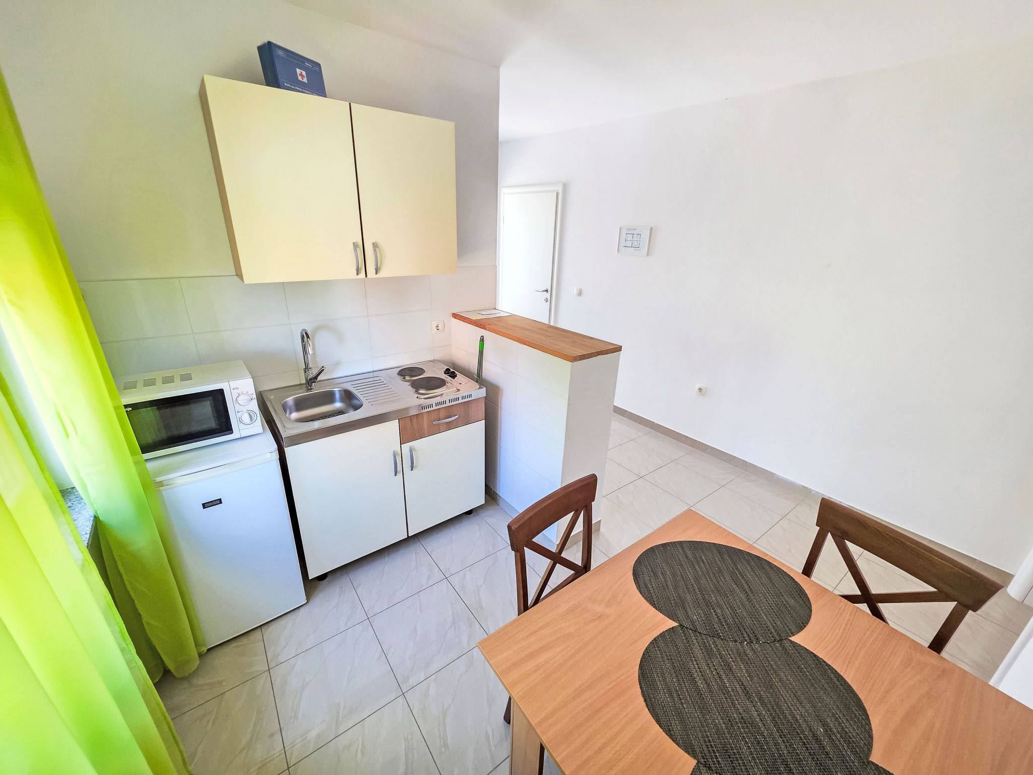 Una cocina compacta con microondas, lavamanos y un refrigerador pequeño. Hay una mesa de madera con un mantel tejido y dos sillas.