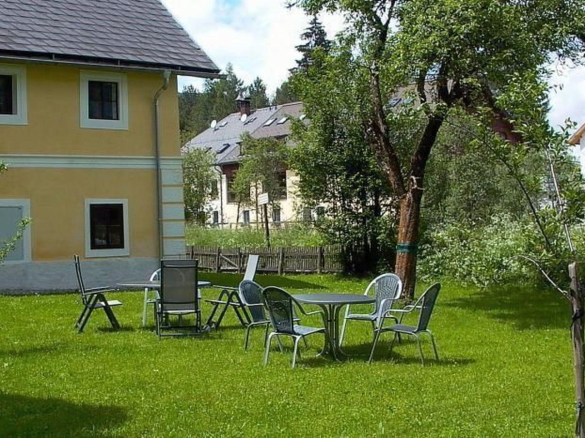 A yellow building with a sloped roof, a grassy yard with a table and chairs, and a tree in the background.