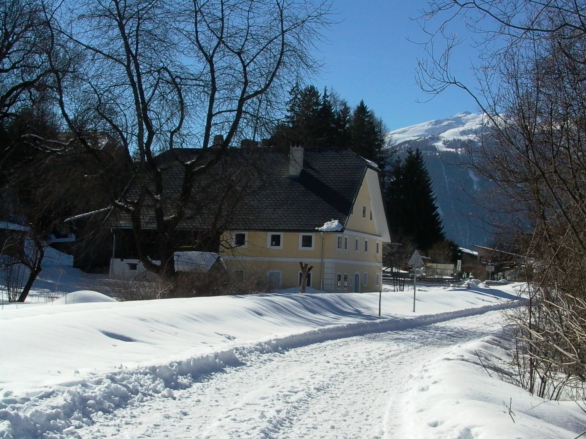 Una carretera rural cubierta de nieve que conduce a una casa amarilla con techo oscuro, rodeada de árboles desnudos y montañas al fondo.