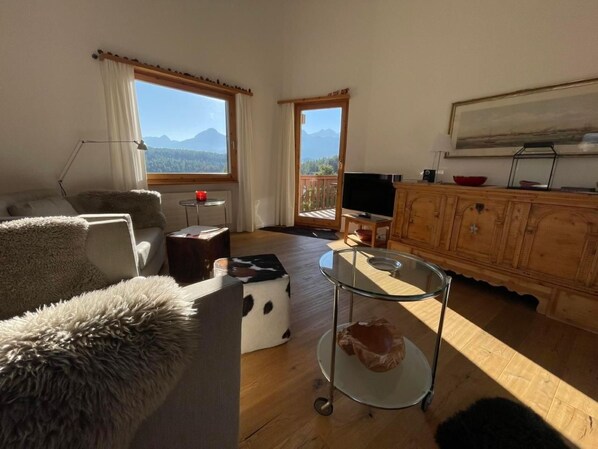 Un salon chaleureux avec un canapé, une table basse en verre et un téléviseur. Il y a des fenêtres donnant sur les montagnes et un meuble en bois.