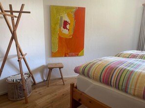 Une pièce avec un lit en bois, un coussin rayé aux couleurs vives, un petit tabouret, un chevalet en bois et un tableau sur toile accroché au mur.