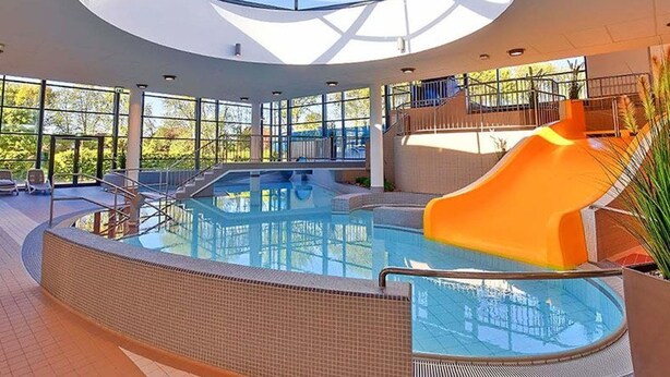 Una piscina interior moderna con techo de vidrio, tobogán y vistas al exterior.
