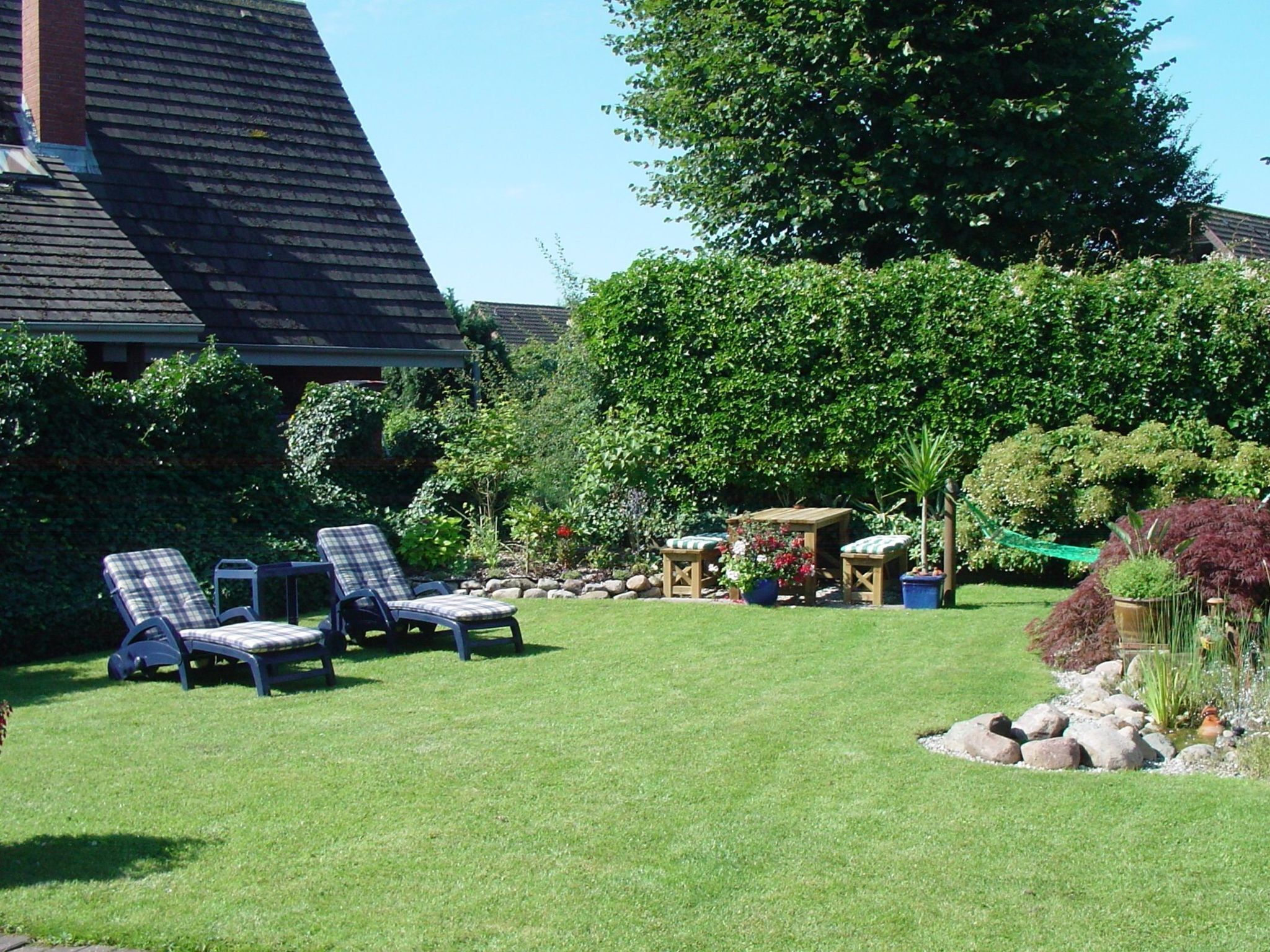 A well-maintained garden with a lawn, two lounge chairs, a wooden table, and a variety of plants and trees.
