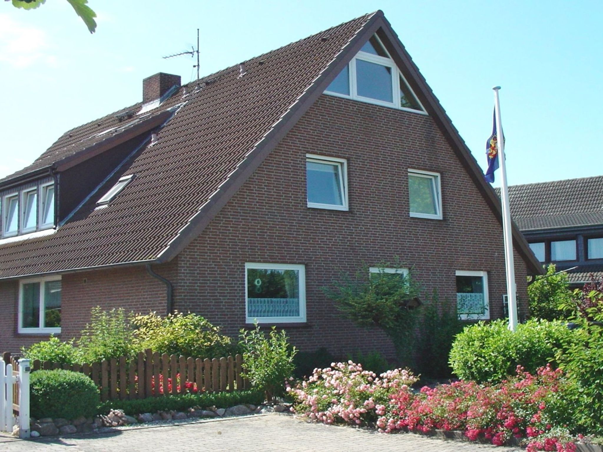 Une maison à deux étages, construite en briques, avec un toit en pente, un mât pour drapeau et une clôture en bois.