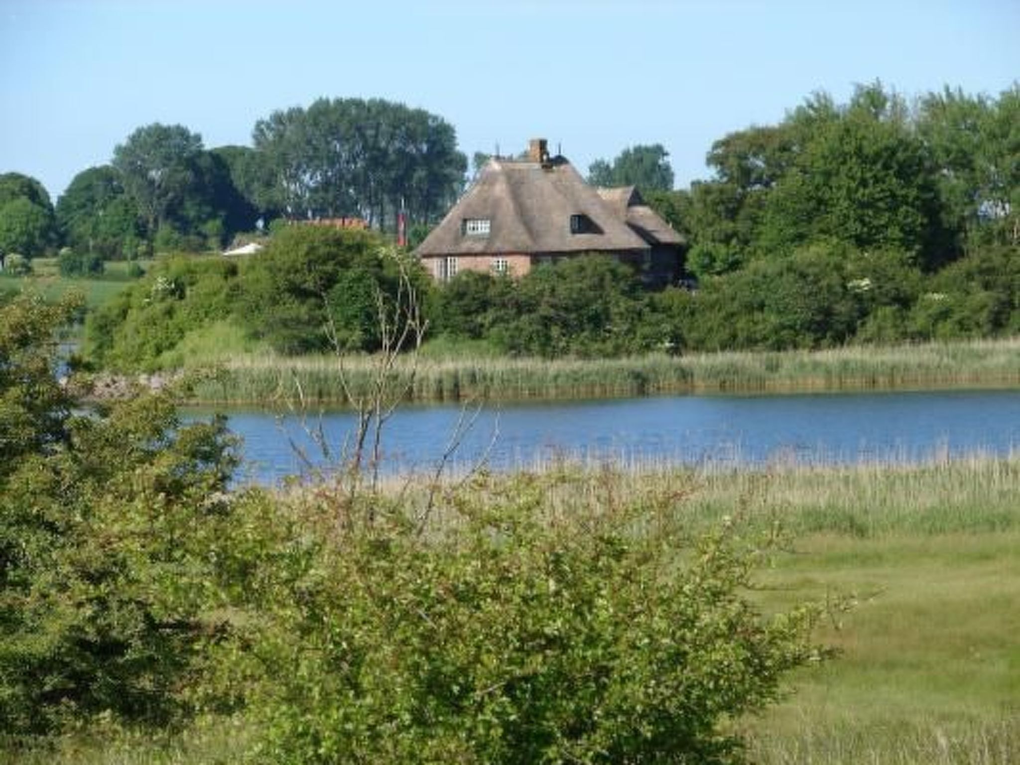 Uma casa com telhado de palha, um lago em primeiro plano e árvores ao fundo.