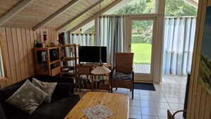 Ett mysigt vardagsrum med ett träpanelat tak, en tv, en bokhylla, en fåtölj i rotting och utsikt över en trädgård genom en glasdörr.