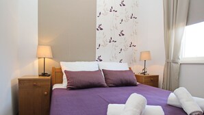 Un letto rifatto con lenzuola viola e due cuscini, un comodino in legno con una lampada, e uno sfondo floreale sul muro.